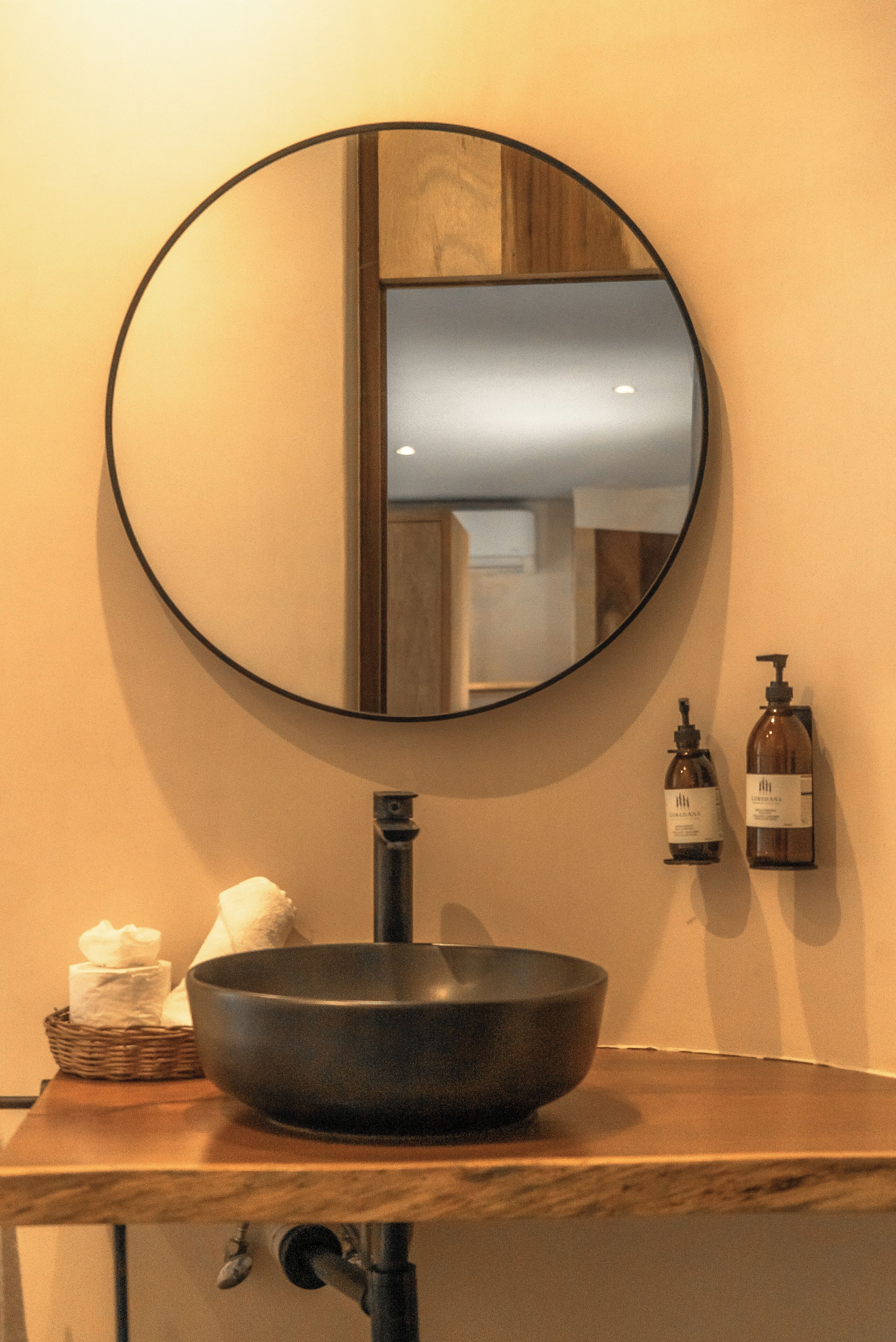 Un baño con un espejo redondo, un lavabo negro, una toalla y dos botellas de jabón líquido en la pared.