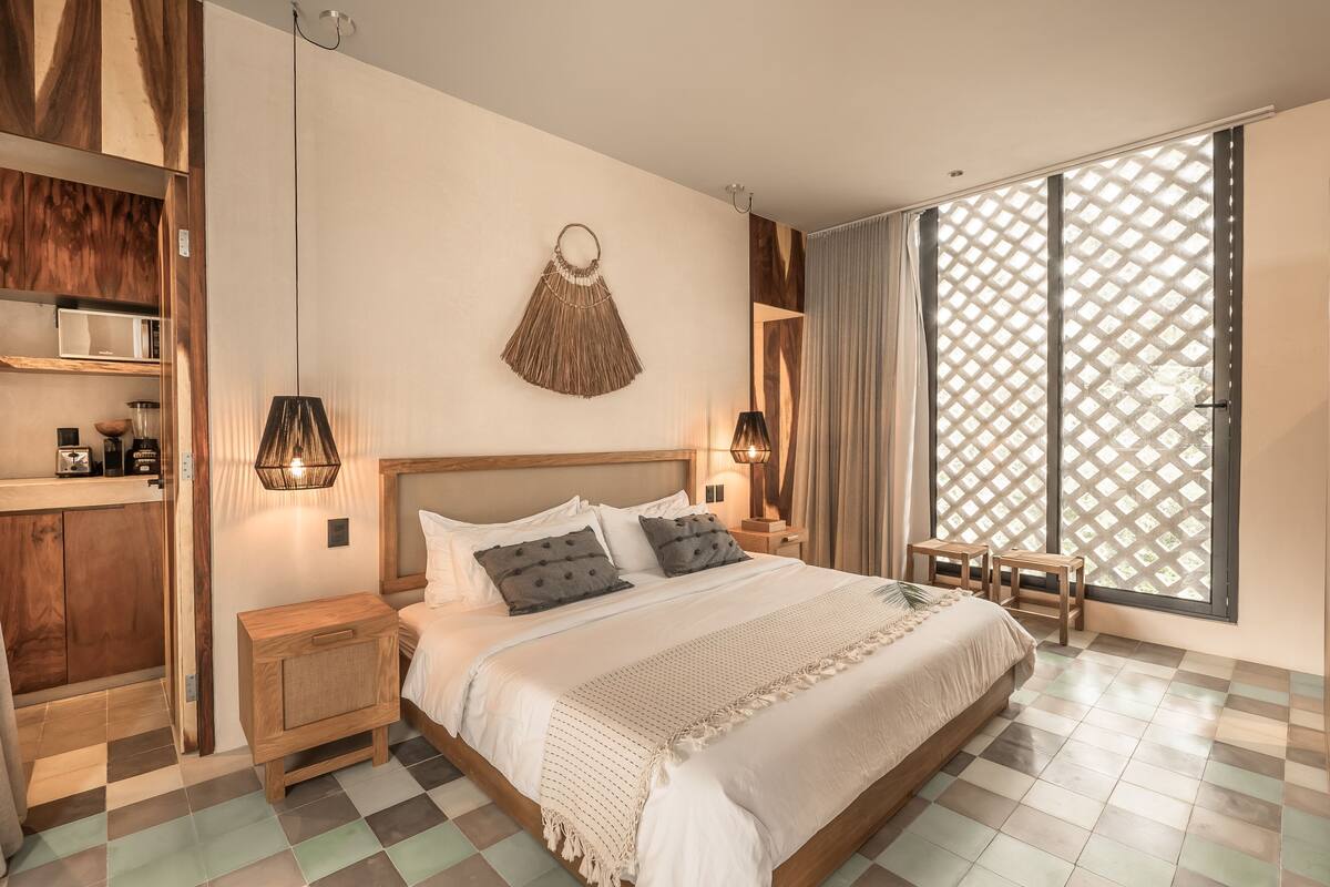 A modern bedroom with a large bed, wooden headboard, bedside tables, a hanging macrame wall decoration, and a large window with a lattice design.