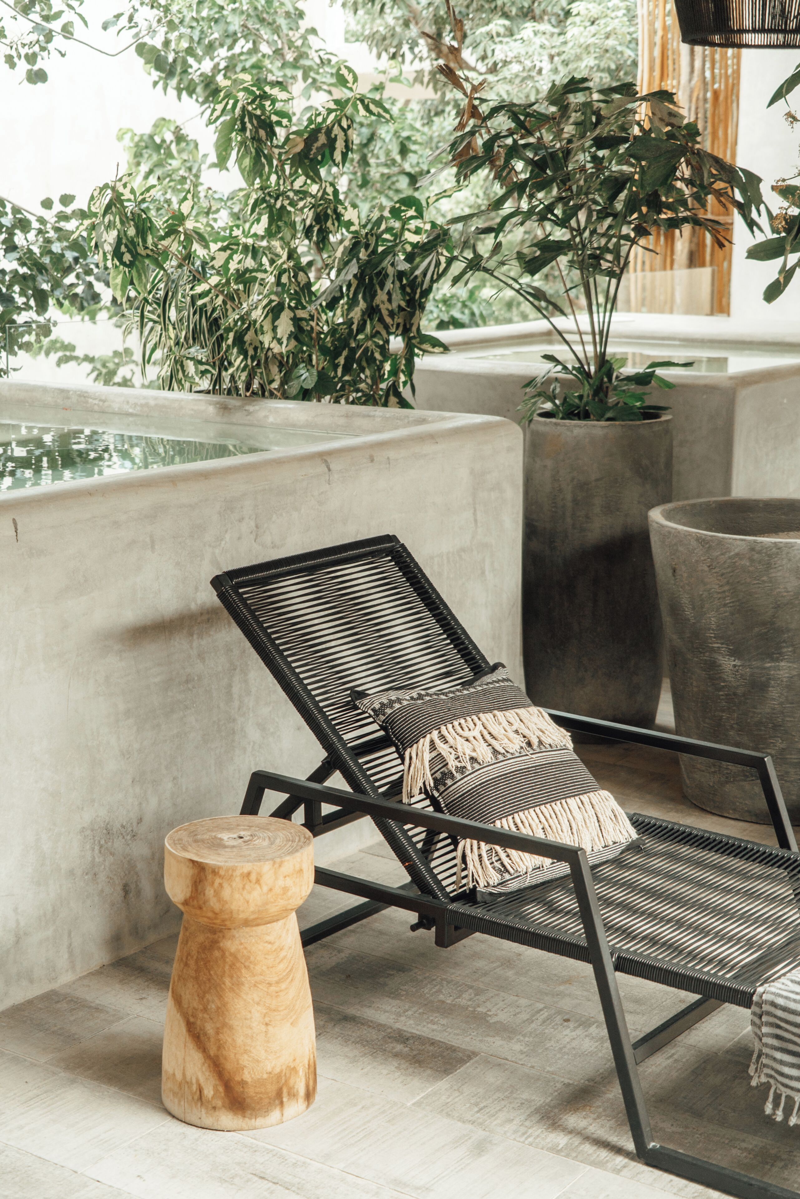 A lounge area with a black metal chair, a wooden stool, and large potted plants.