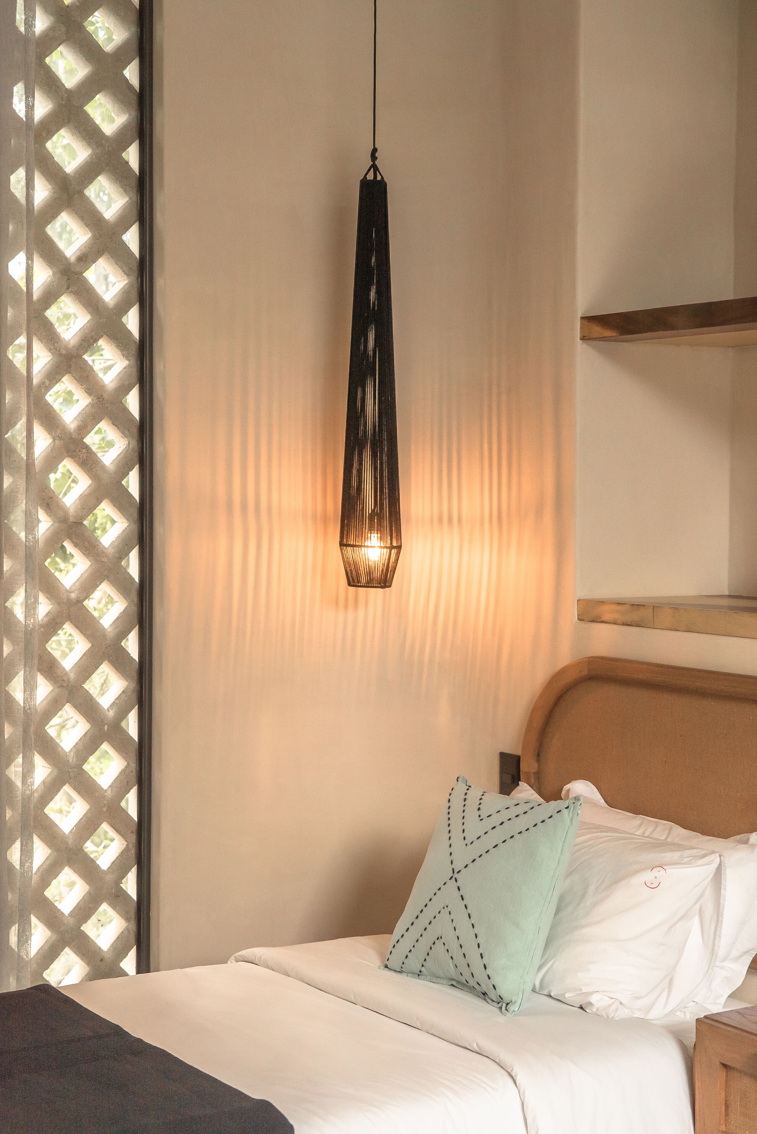 A neatly made bed with a light-colored headboard, a hanging pendant light, and a lattice window to the left.
