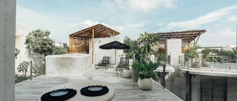 An outdoor patio with a round table, chairs, and umbrellas, surrounded by potted plants and a pool.