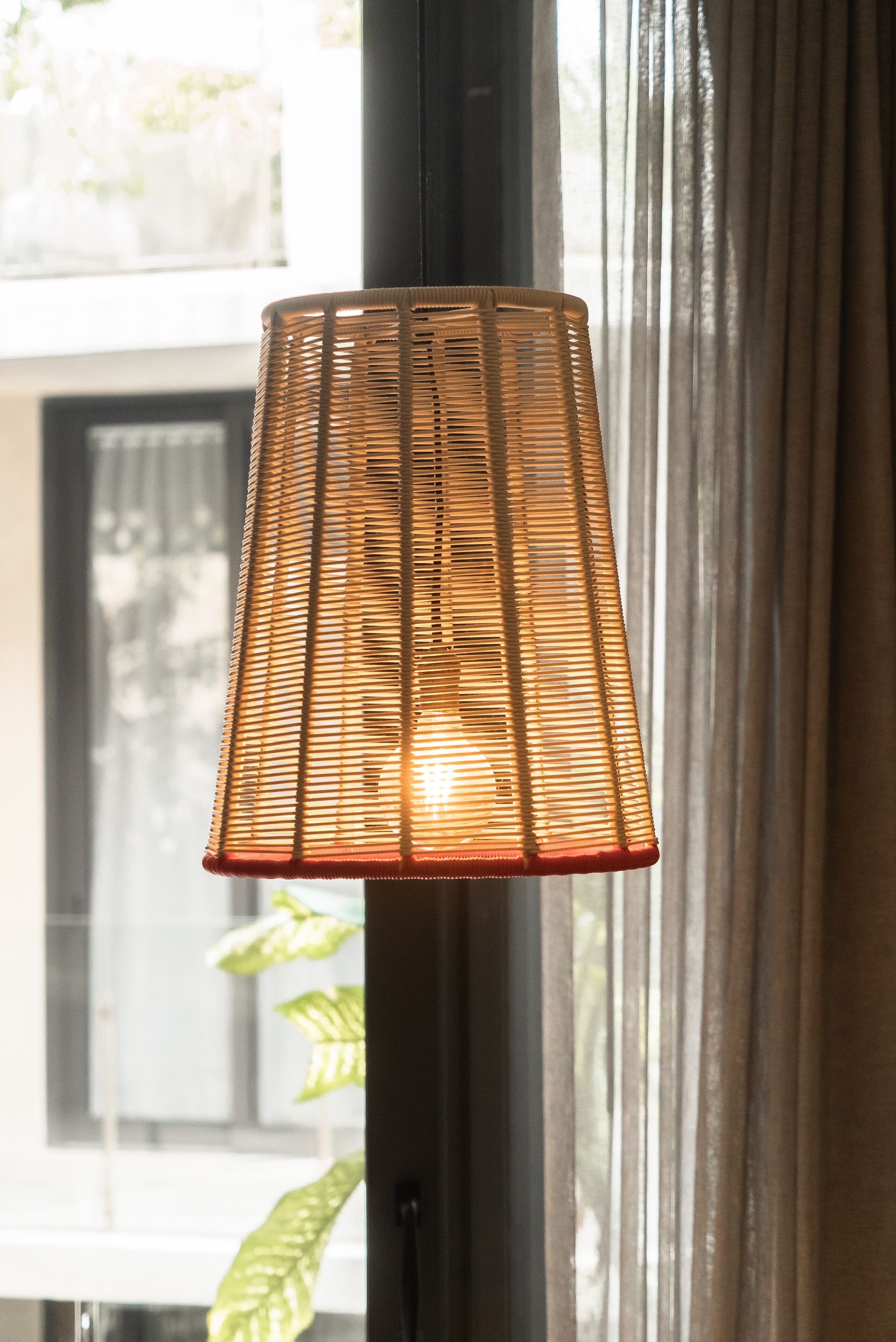 Una lámpara de mimbre con base roja, colgando cerca de una ventana con cortinas.