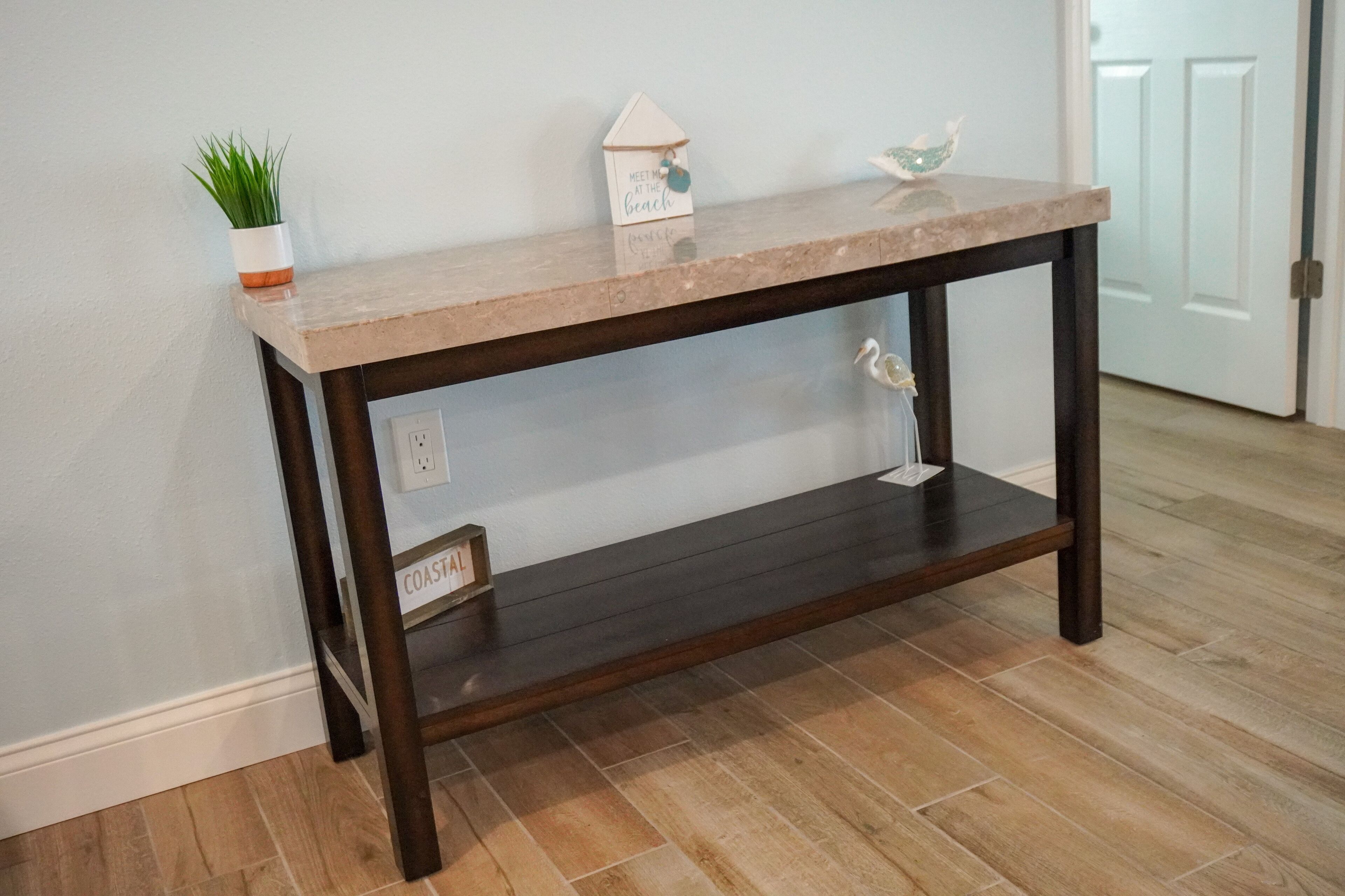 Une console en bois foncé, surmontée d’un plateau de couleur claire, agrémentée d’une petite plante en pot, d’un panneau décoratif en forme de maison et d’un ornement en verre.