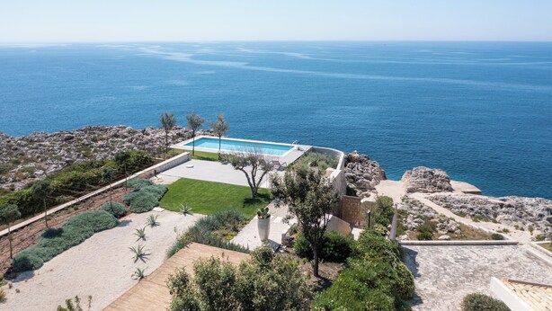 A pool with a view of the sea, surrounded by landscaped garden and rocky terrain.