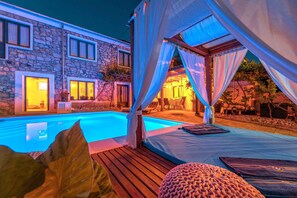 Un espace aménagé au bord de la piscine, avec une terrasse en bois, un coin salon couvert doté de coussins et, au fond, un bâtiment en pierre.