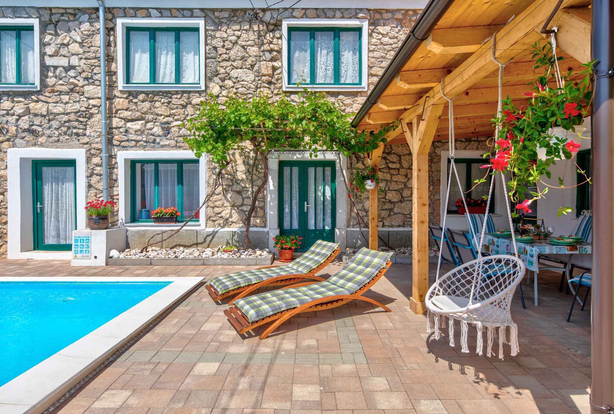 Un bâtiment en pierre doté d’une piscine, de deux chaises longues et d’un fauteuil suspendu.