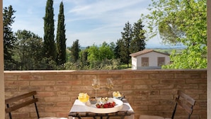 Eine Terrasse mit Tisch für zwei Personen, mit Erdbeeren auf einem Teller, einer Oliven-Schale und zwei Gläsern Weißwein. Die Terrasse bietet einen Ausblick auf eine reizvolle Landschaft mit Bäumen und einer entfernten Ansicht eines kleinen Gebäudes.
