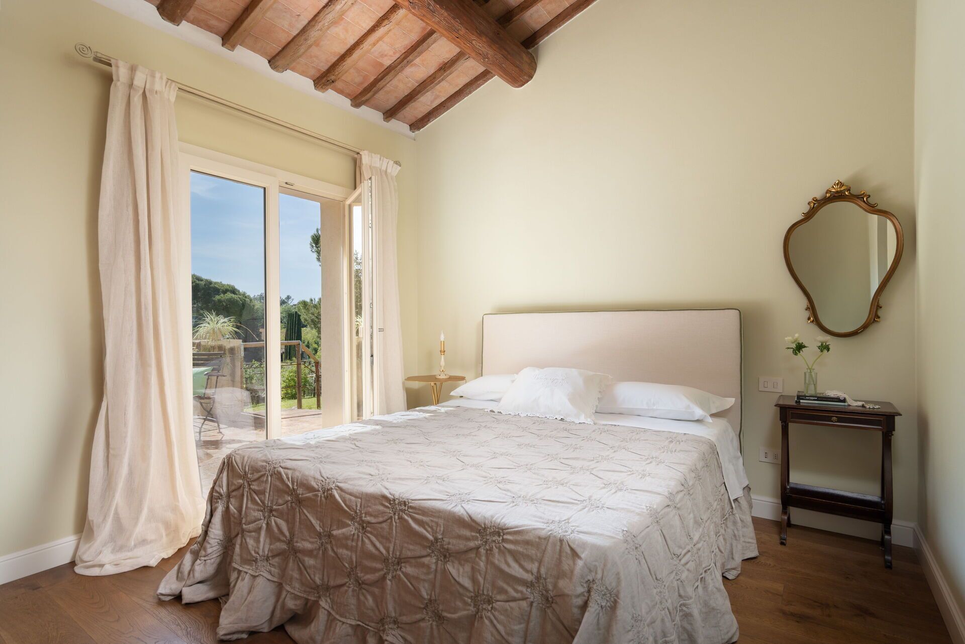 A bedroom with a large bed, a wooden ceiling, a window with curtains, a bedside table with a lamp, and a mirror on the wall.