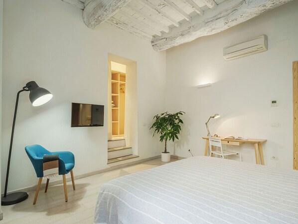 A bedroom with a bed, a desk with a chair, a blue chair, a floor lamp, a potted plant, and a TV on the wall. - Apartment in Lucca with 1 bedrooms sleeps 2 (Lucca)