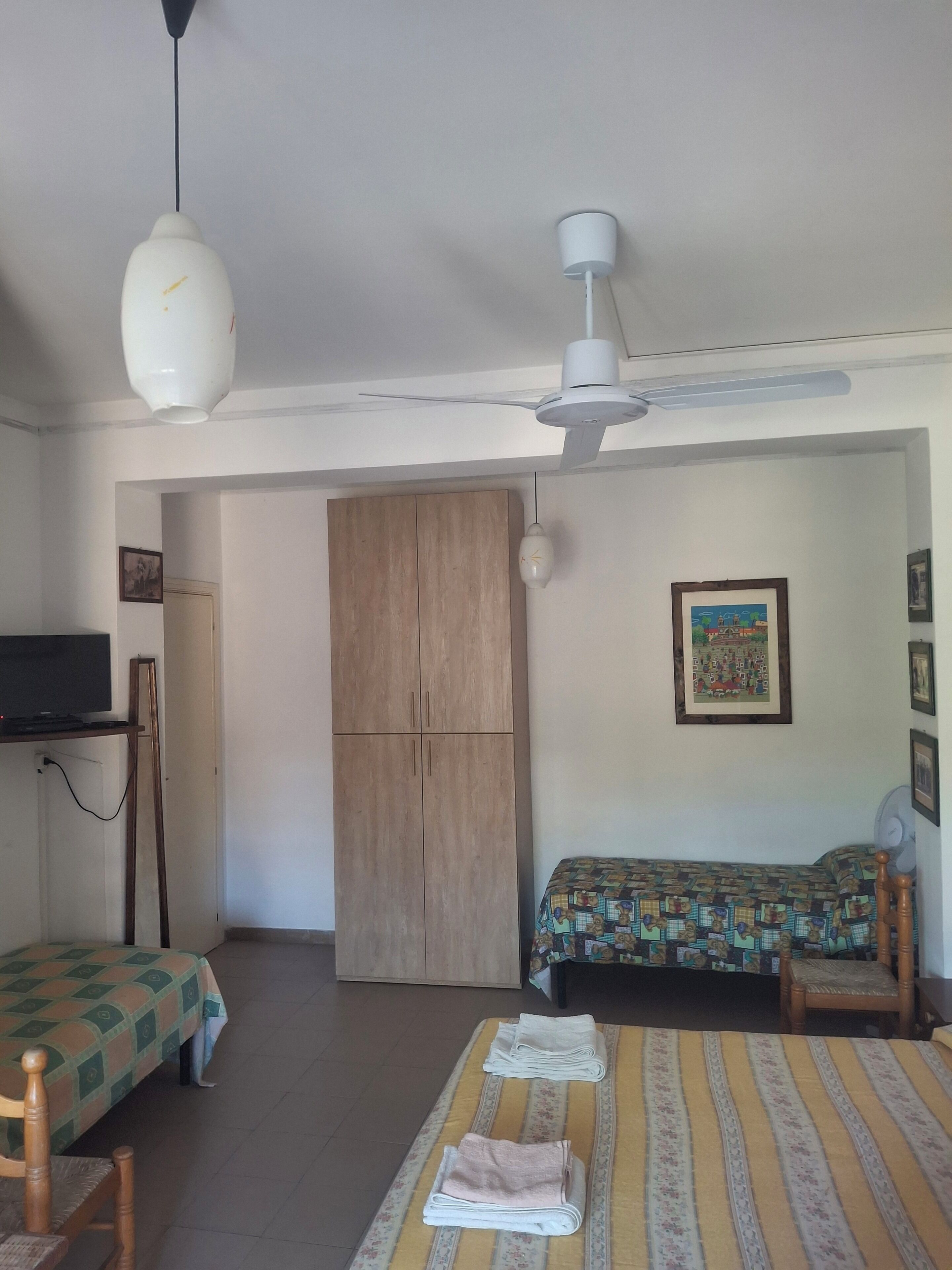 Una stanza con un letto, una scrivania con un computer, una sedia, un armadio e una ventola a soffitto.
