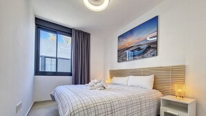 Ein Hotelzimmer mit einem großen Bett, einem Nachttisch, einem Fenster mit Vorhängen und einem Gemälde von einem Sonnenuntergang über einem Strand.