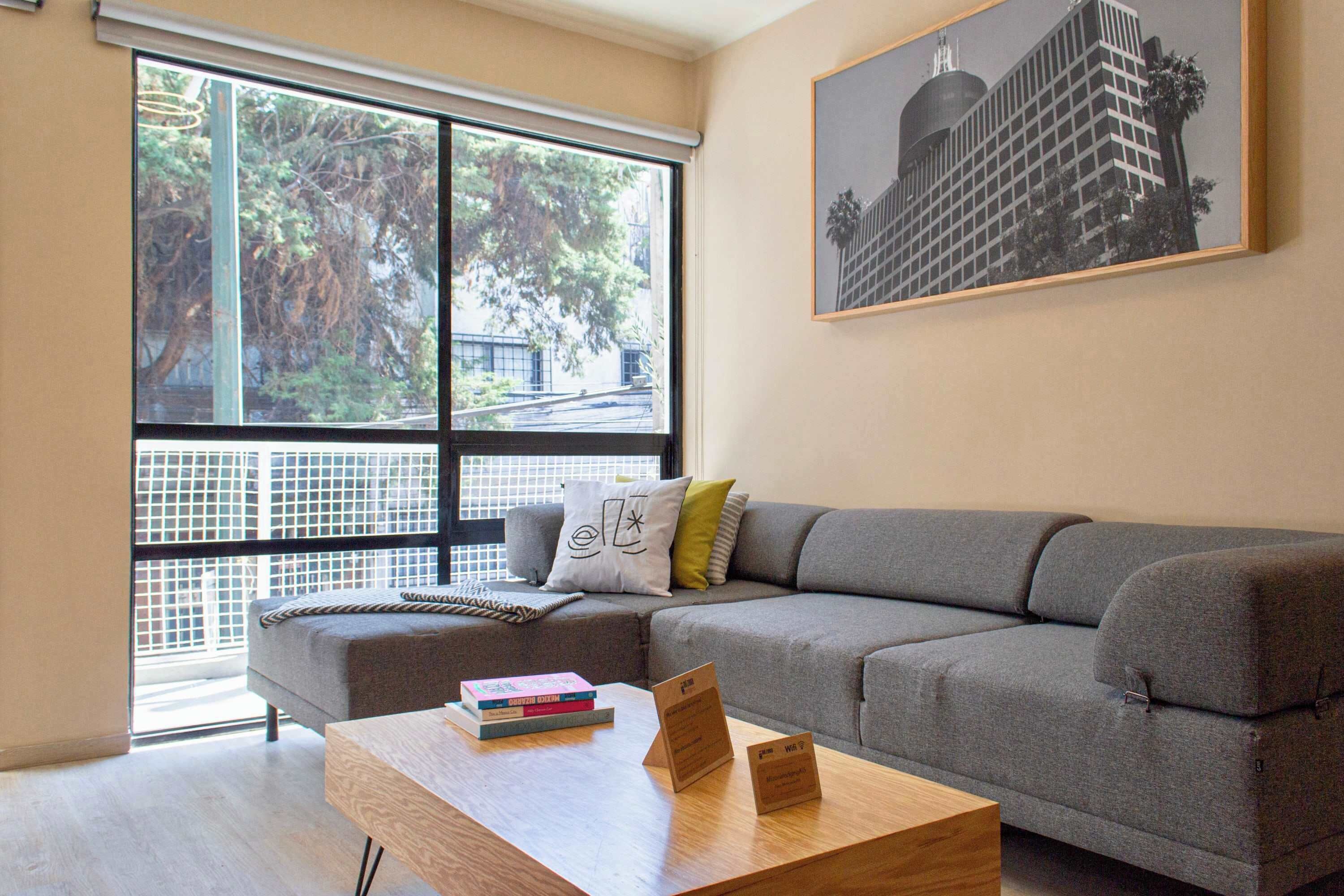 A modern living room with a grey sofa, a wooden coffee table with books, and a large window with a view of trees and buildings.