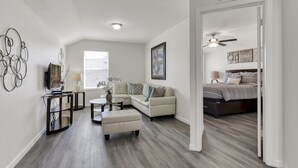 Un salon moderne avec un canapé d’angle, une table basse et un téléviseur. Il y a une chambre avec un lit et un ventilateur de plafond. La pièce est dotée d’un plancher en bois franc et de murs blancs.