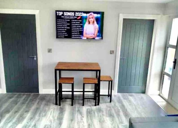 Quarto com mesa de madeira e duas cadeiras, uma TV na parede exibindo 'TOP SONGS 2022', e uma porta com painel azul.