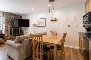 A cozy living space with a stone wall, a flat-screen TV, a wooden dining table with chairs, a sofa, and a kitchen area with a dishwasher.