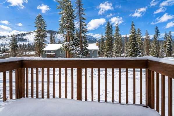 A wooden deck with a snow-covered ground, surrounded by snow-laden trees and a mountain backdrop.
