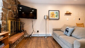A living room with a stone wall, a flat-screen TV mounted above a fireplace, a framed picture on the wall, a wooden floor, and a grey sofa with a decorative pillow.