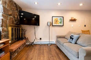 Ein Wohnzimmer mit Steinwand, einem Flachbildfernseher über einem Kamin, einem gerahmten Bild an der Wand, Holzboden und einem grauen Sofa mit Zierkissen.