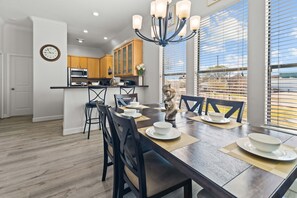 A modern kitchen with wooden cabinets, a dining area with a wooden table set for four, and a large window with blinds.