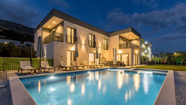 Una casa moderna de dos pisos con piscina, sillas de descanso e iluminación exterior.