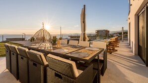 Un área de comedor al aire libre con vista al mar, una pérgola y un edificio moderno al fondo.