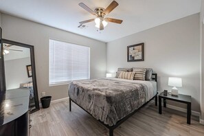 A bedroom with a bed, a ceiling fan, a window with blinds, a bedside table with a lamp, and a dresser.