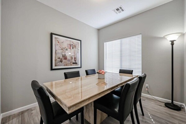 A dining area with a marble table, black chairs, a framed abstract painting, a window with blinds, and a floor lamp.