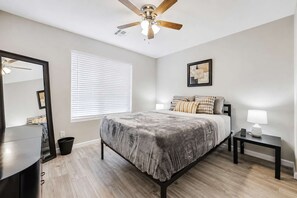 A bedroom with a bed, a ceiling fan, a bedside table with a lamp, a mirror, and a window with blinds. - Pet-Friendly 3BR | Fenced Yard | King Bed (Chandler)