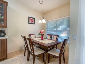 Un comedor con mesa de madera, seis sillas, un florero con flores, una lámpara colgante y una ventana con persianas.