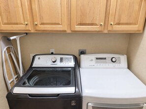 A laundry area with a front-loading washer and dryer, wooden cabinets above, and a metal drying rack to the left. - Pet-Friendly 3BR | Fenced Yard | Lake Trails (Tempe)