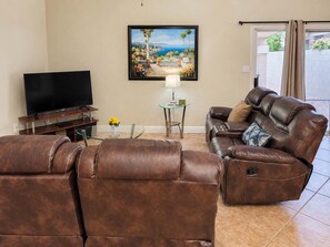 A living room with a leather sofa, a flat-screen TV, a glass coffee table, and a painting of a coastal scene.
