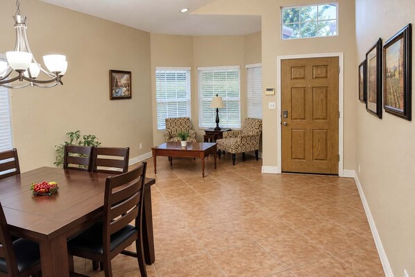 A dining area with a wooden table, chairs, and a chandelier. There are two windows with blinds, a door, and framed pictures on the wall.