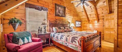 A cozy bedroom with a wooden ceiling, a bed with a patterned quilt, a red armchair, a nightstand, a lamp, and a ceiling fan.