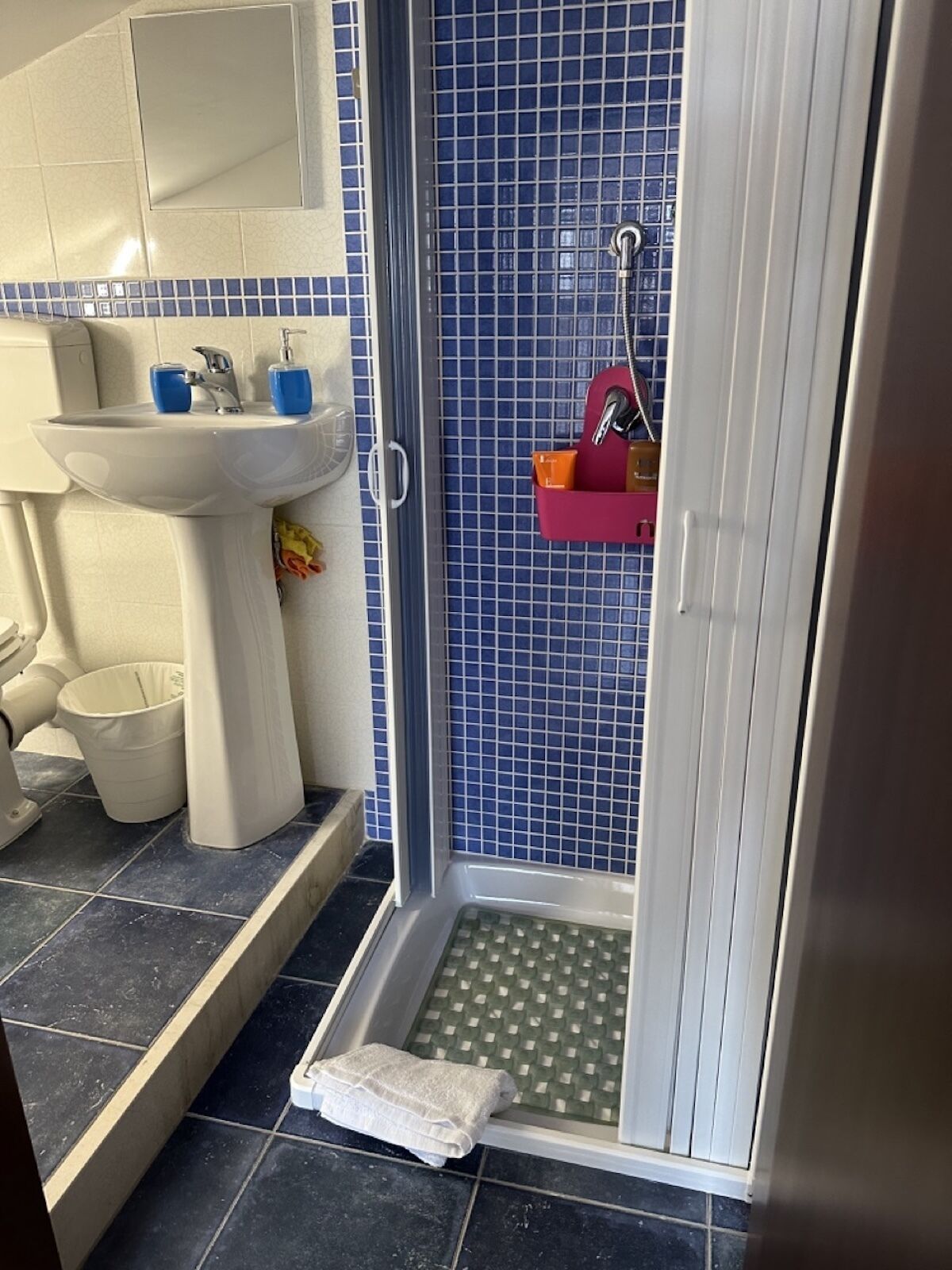 A bathroom with a white sink, blue-tiled shower, and a white towel on the floor.