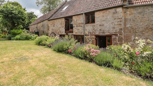 Un bâtiment en pierre avec un toit en tuiles, un jardin avec différentes plantes et fleurs, et une brouette au premier plan.