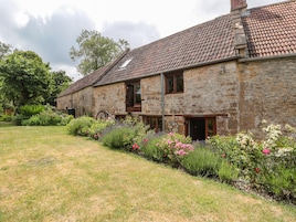 A stone building with a tiled roof, a garden with various plants and flowers, and a wheelbarrow in the foreground.