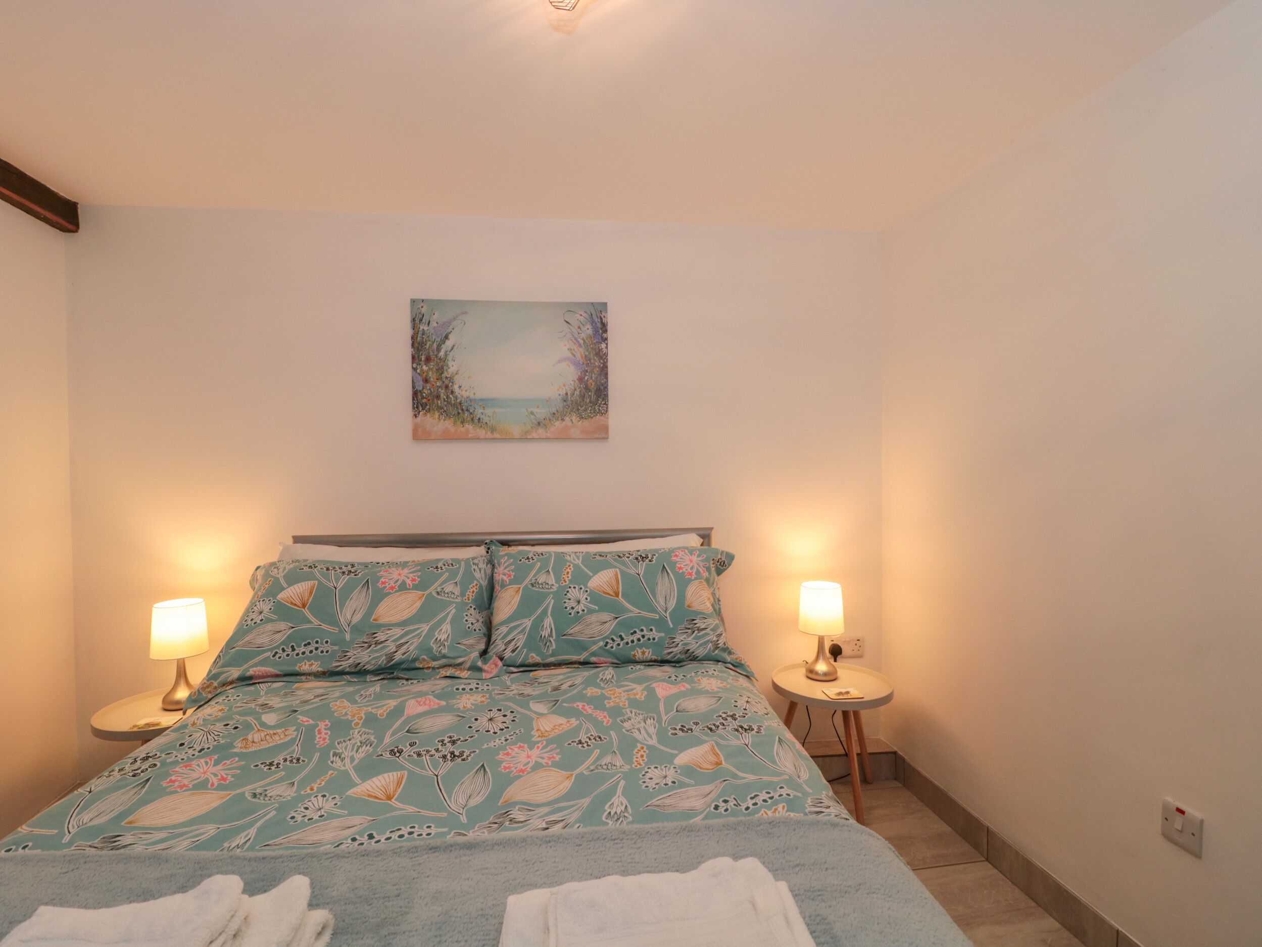 A bedroom with a bed, two bedside lamps, a painting on the wall, and a small table with a lamp.