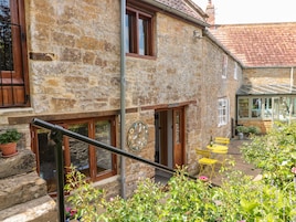 A stone building with a wooden door labeled "The Old Grange," a small outdoor seating area with yellow chairs, and a staircase leading to the entrance.