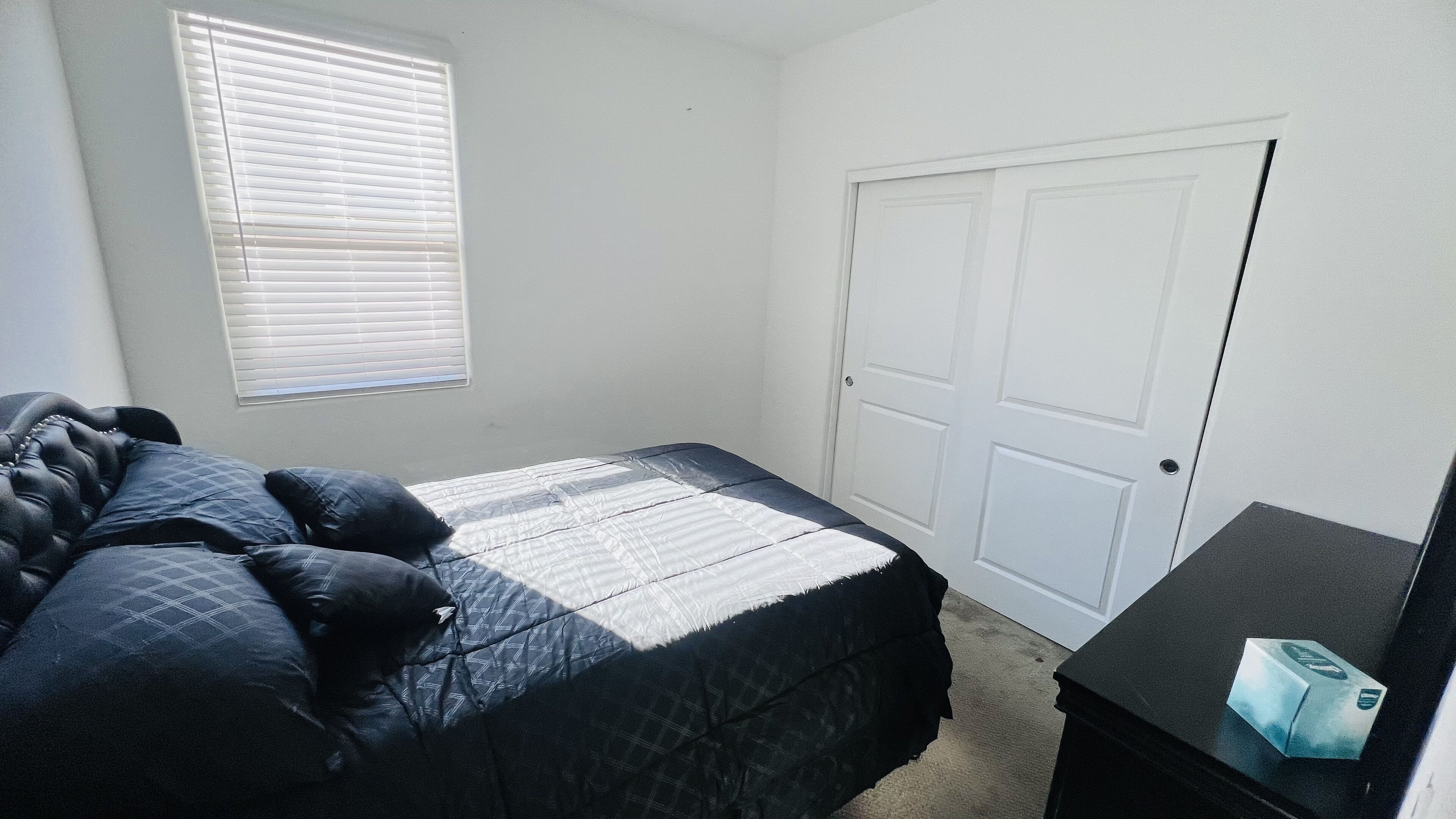Une chambre à coucher avec un lit, une fenêtre avec des stores, une porte et une petite table sur laquelle se trouve une boîte.