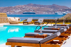 Un área junto a la piscina con sillones de descanso y una vista despejada del mar y las montañas al fondo.