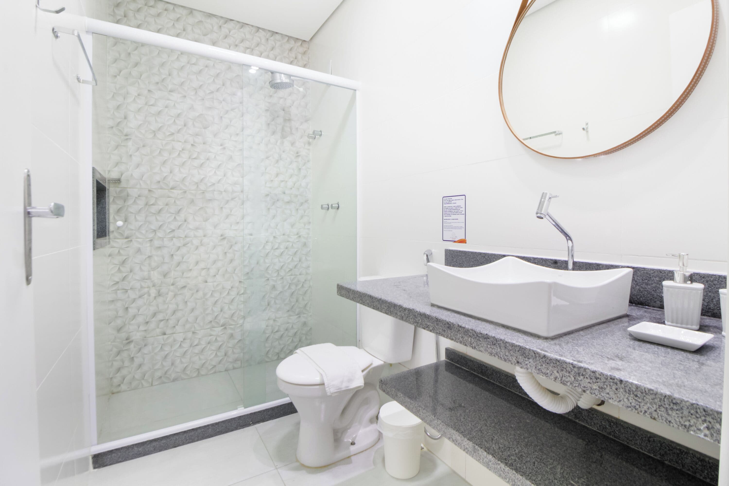 A modern bathroom with a glass-enclosed shower, a white toilet, a granite countertop, and a round mirror.