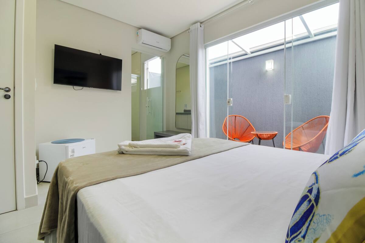 A hotel room with a bed, a TV mounted on the wall, an air conditioning unit, a bathroom visible through a glass door, and two orange chairs outside.