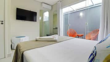 A hotel room with a bed, a TV mounted on the wall, an air conditioning unit, a bathroom visible through a glass door, and two orange chairs outside.