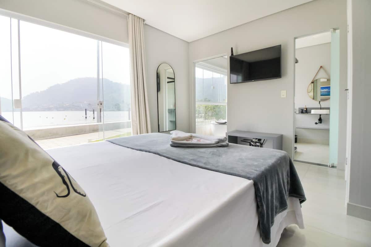 A modern hotel room with a large bed, a flat-screen TV, a bathroom with a mirror, and a view of the sea.