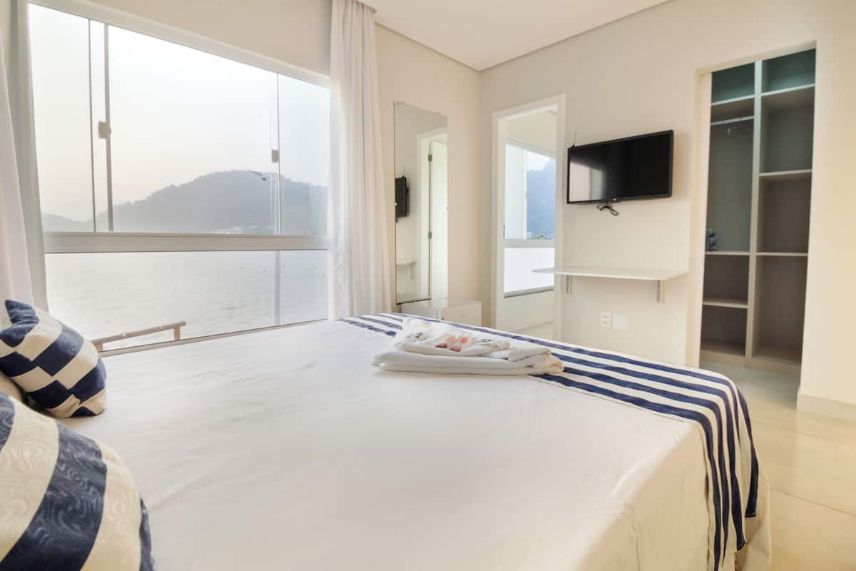 A hotel room with a large bed, a TV mounted on the wall, and a view of the sea through a large window.