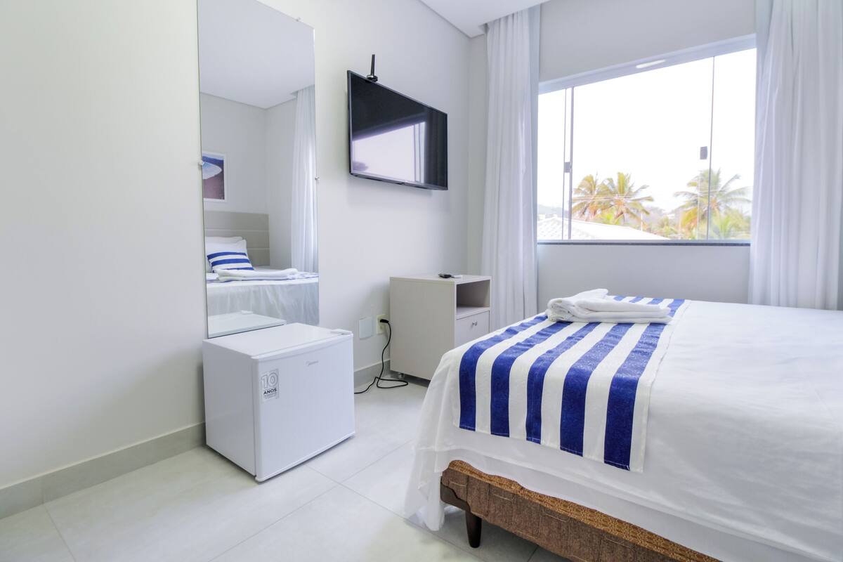 A hotel room with a bed, a small white refrigerator, a mirror, and a window with a view of palm trees.