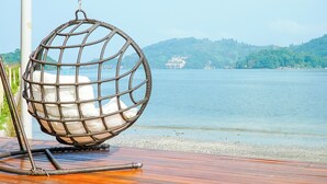 A wicker hanging chair on a wooden deck overlooking a calm lake with a distant cruise ship and forested hills.