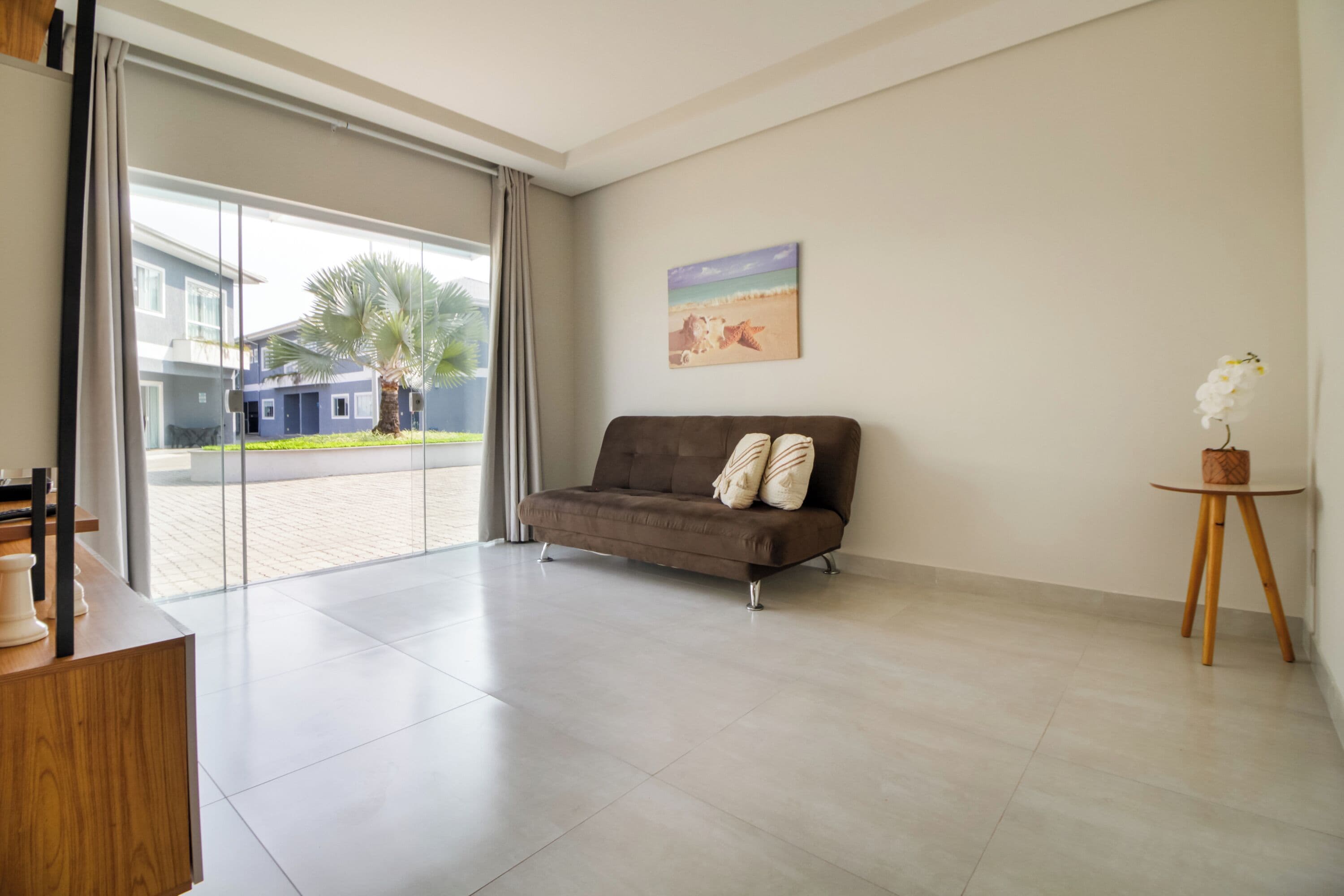 A modern living room with a brown sofa, a small round table with a white orchid, and a painting of a beach scene.