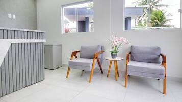 Uma área de recepção moderna com balcão, duas poltronas cinza acolchoadas, uma pequena mesa redonda com planta e amplas janelas com vista para palmeiras.