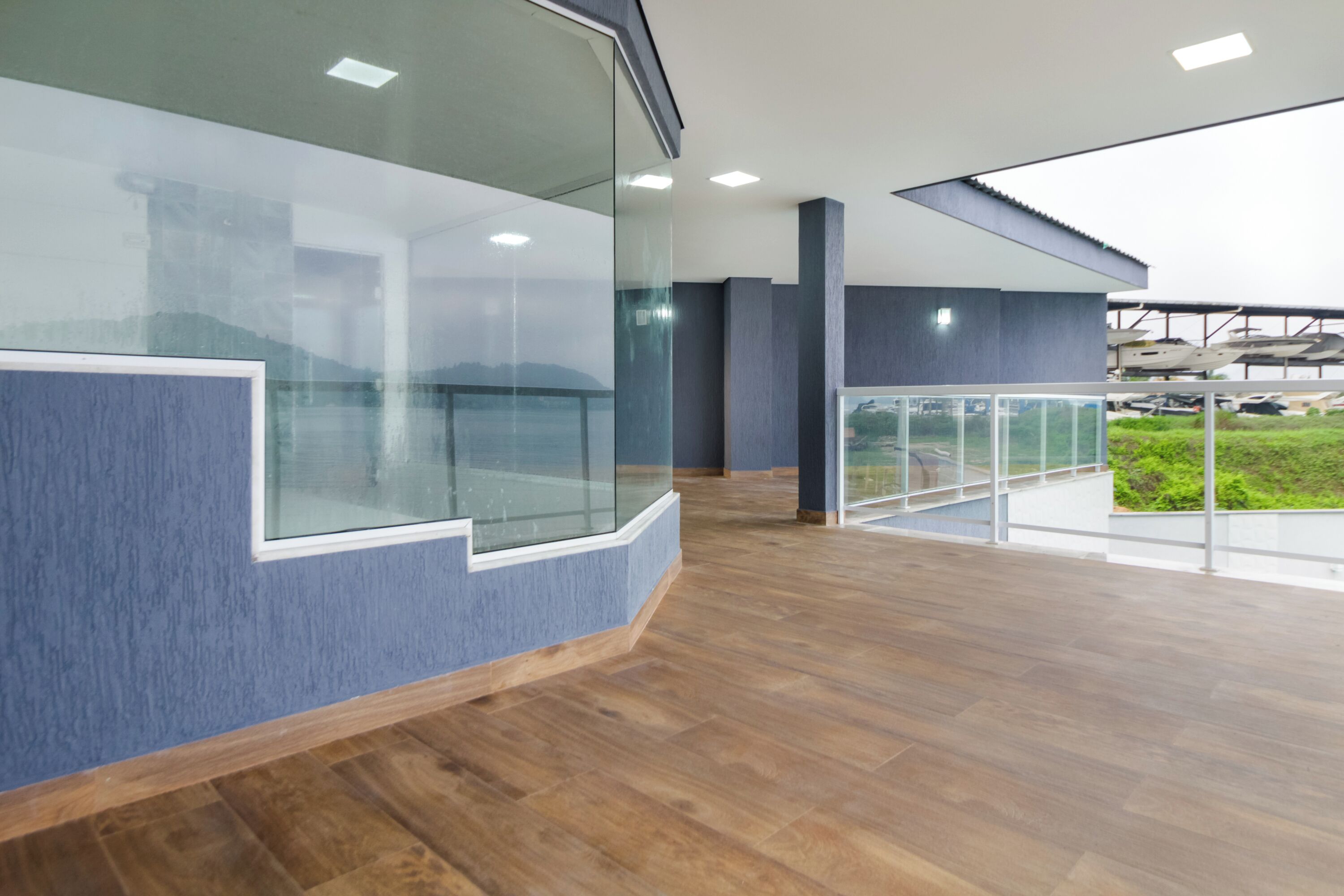 A modern interior with a curved glass partition, wooden flooring, and a view of parked boats through large windows.